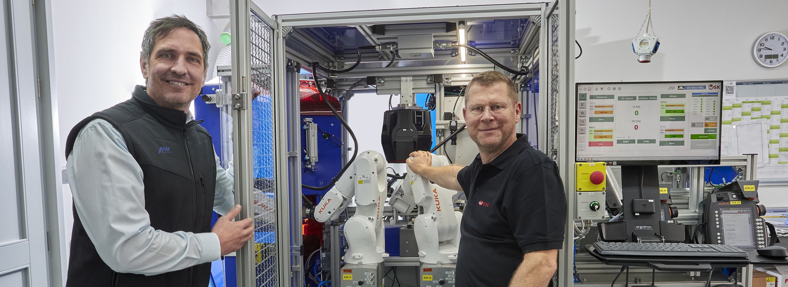 Zwei Mitarbeiter stehen neben einer automatisierten KUKA-Roboteranlage in einer modernen Produktionsumgebung und blicken in die Kamera.